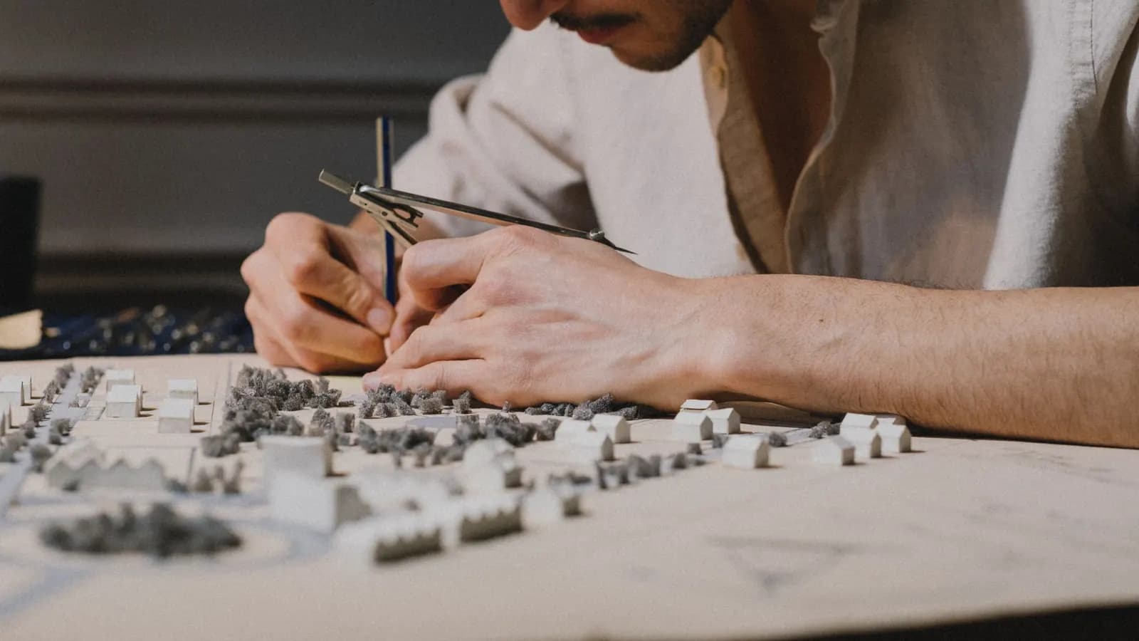 A person leaning over a card site model with small white houses and foam trees, using dividers