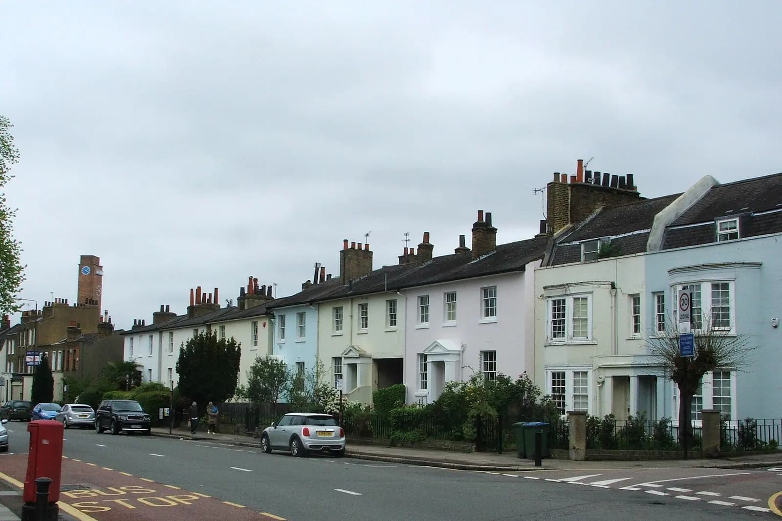 A terrace of pastel-painted Regency villas along Greenwich South Street, with a red post box, a bus stop and a brick clock tower behind