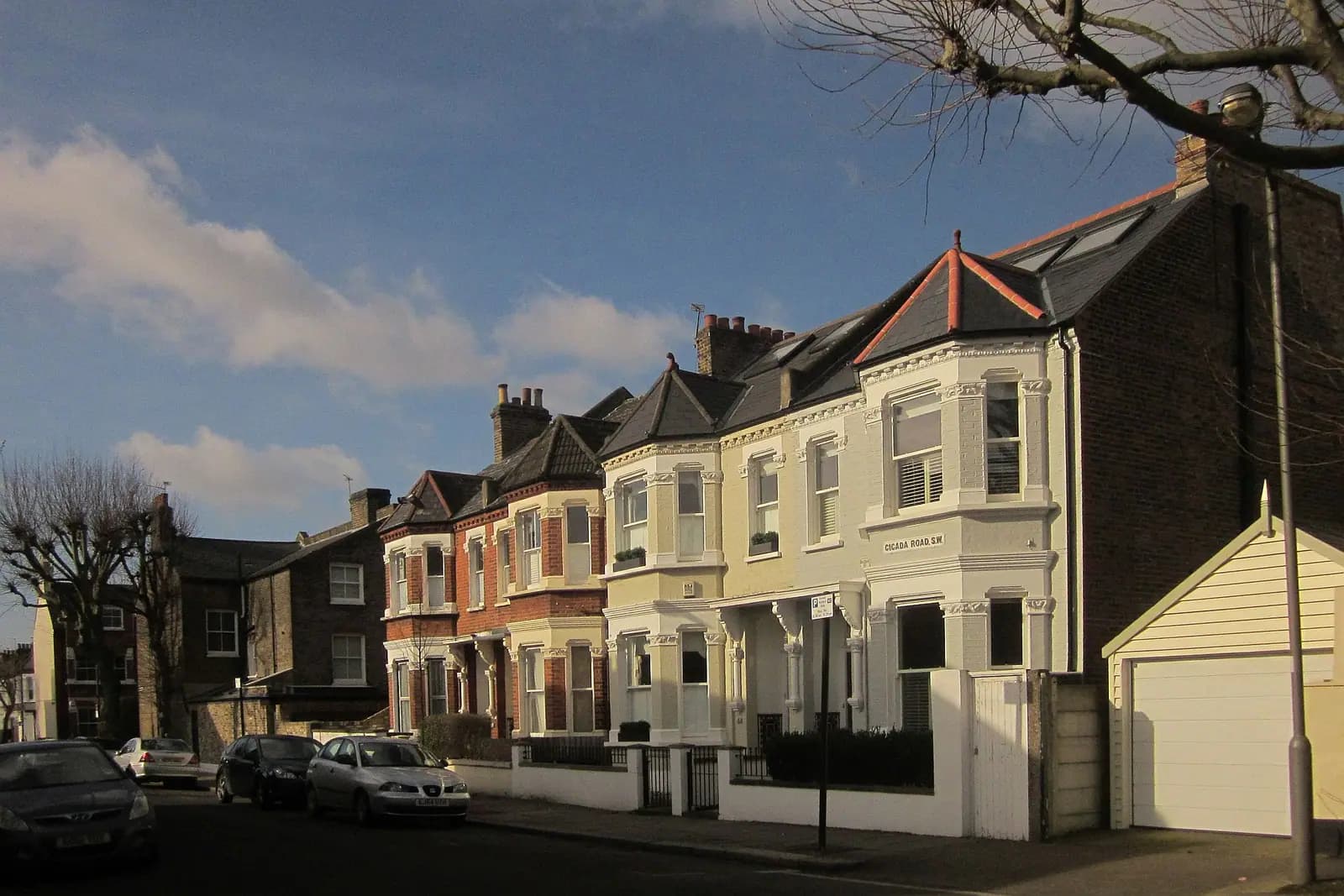 A corner of Victorian houses on Cicada Road in Wandsworth with bay windows, one with a roof dormer, in warm evening light and a garage at the right