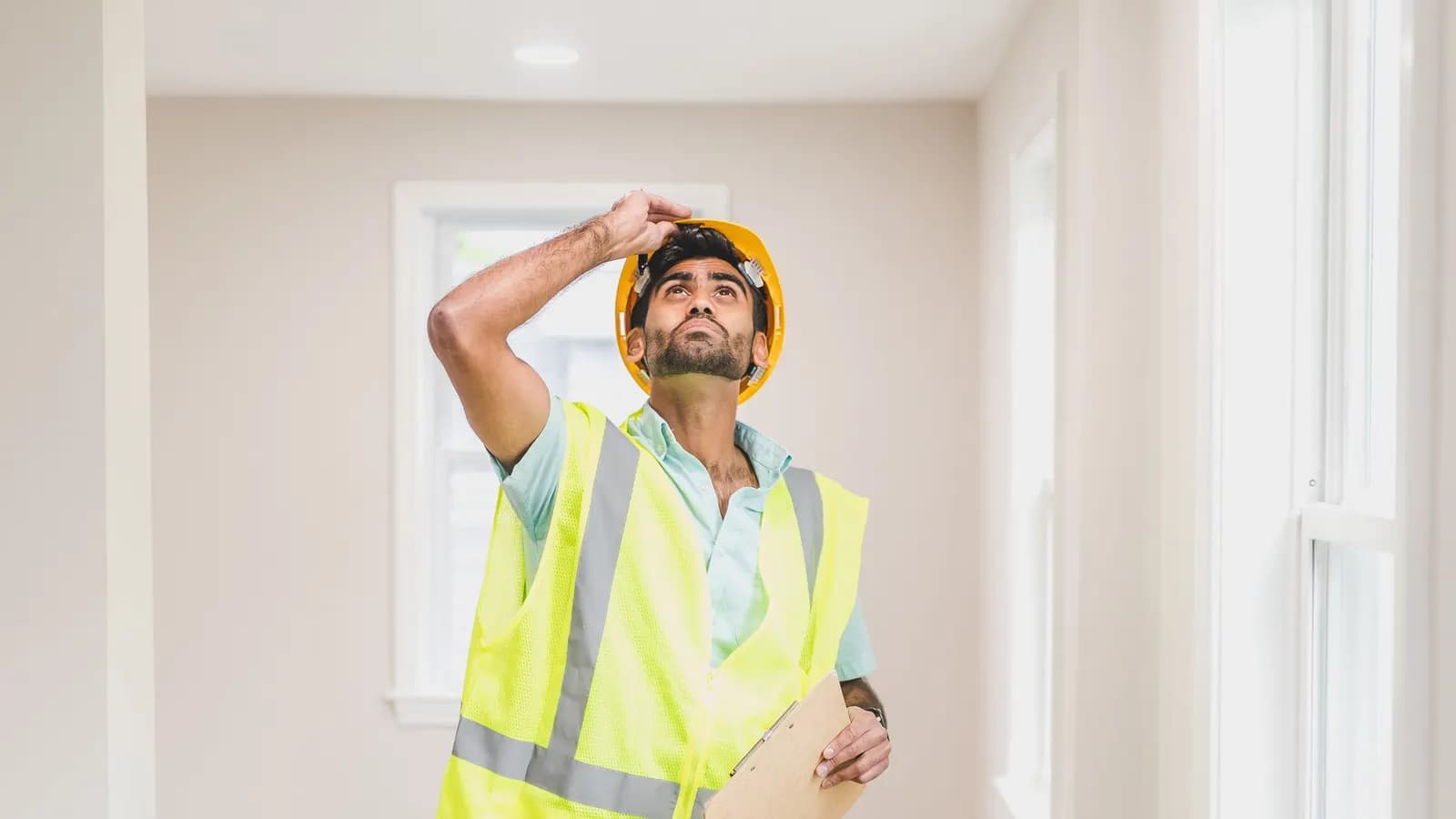 An inspector in a yellow hard hat and high-visibility vest holding a clipboard and looking up at the ceiling of an empty, freshly decorated room