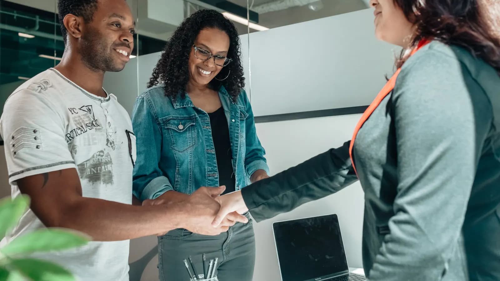 A smiling couple shaking hands with an advisor in a bright office, with a laptop and a pot of pencils behind them