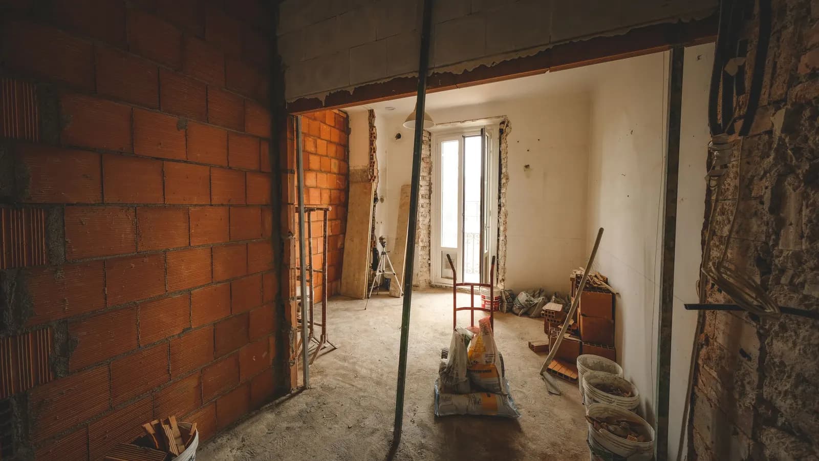 A partly renovated room with bare orange brick walls, a new opening, bags of cement and buckets of debris on the floor