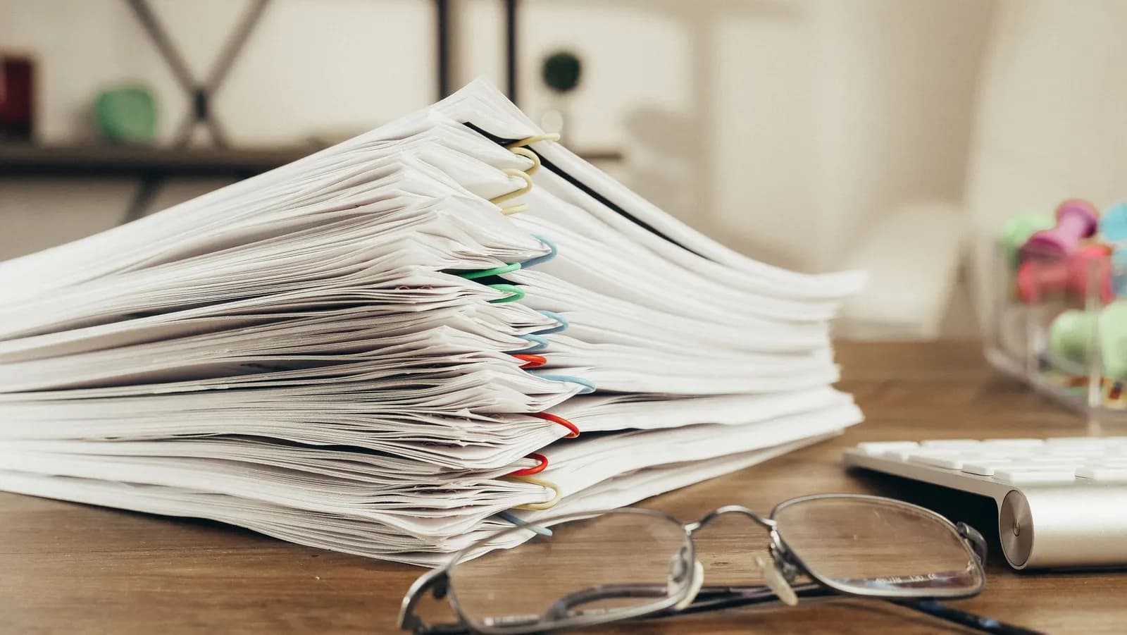 A tall stack of office papers held with coloured clips on a wooden desk, a pair of glasses and a keyboard in front