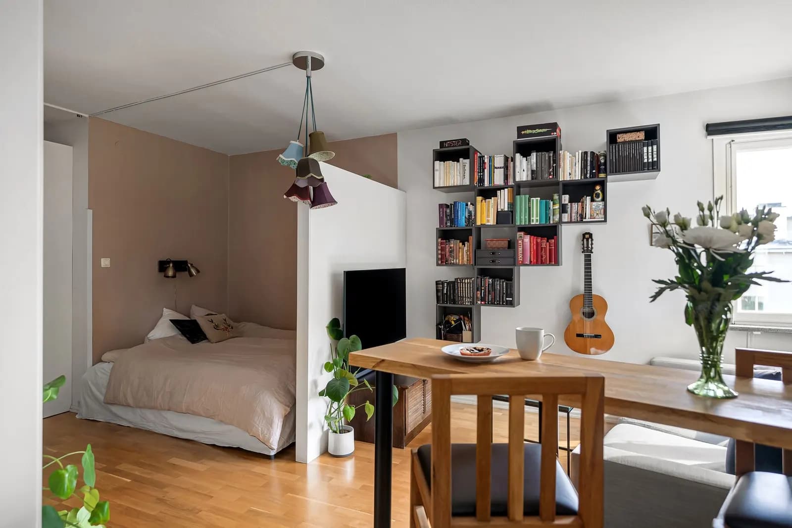 An open-plan studio with a bed in an alcove behind a low partition, a wall of shelves with books, a guitar and a wooden dining table