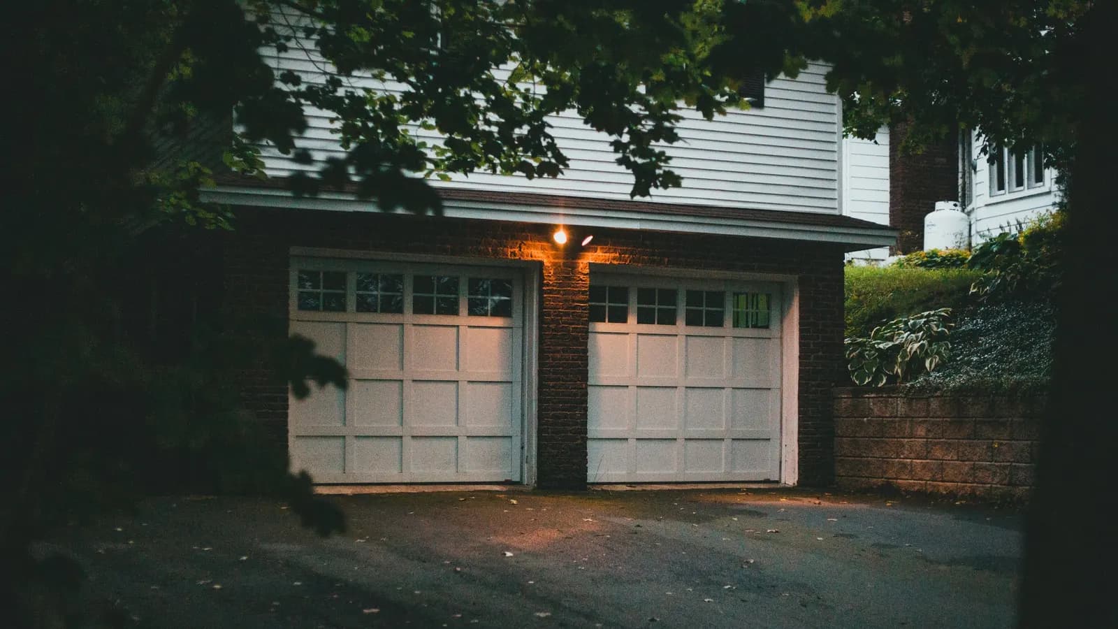 A brick double garage with two white panelled doors lit by one lamp at dusk, under the upper floor of a clapboard house