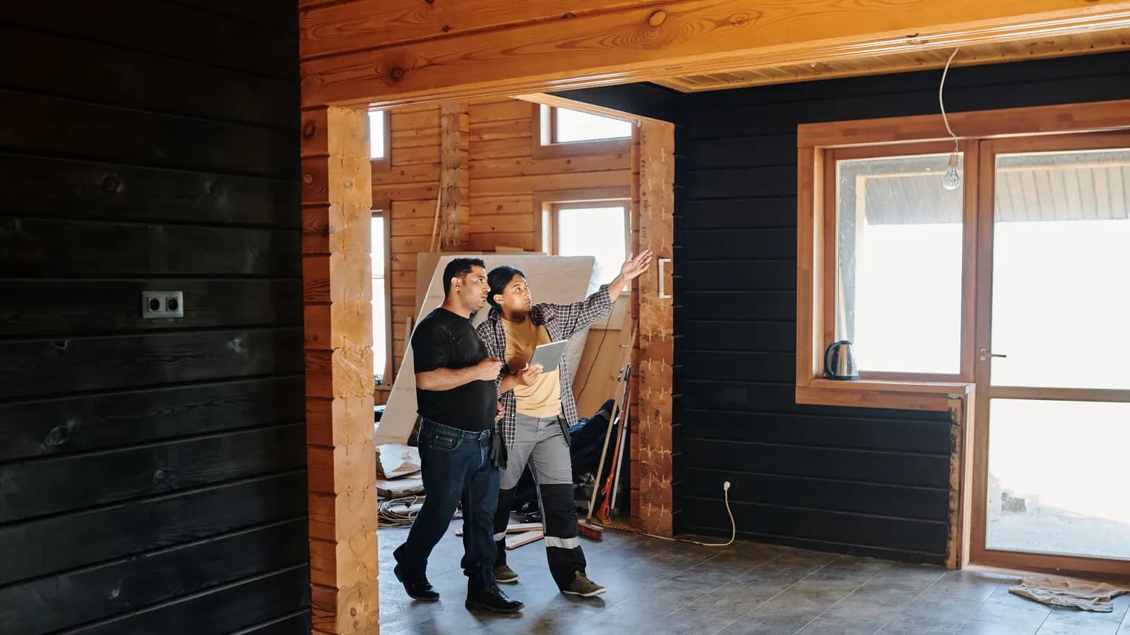Two people walking through an unfinished timber-clad room, one pointing up while holding a tablet