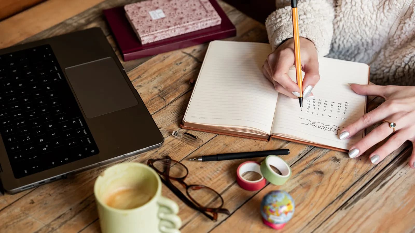 A woman writing in an open notebook with a pencil on a wooden desk beside a laptop, a coffee mug and a pair of glasses