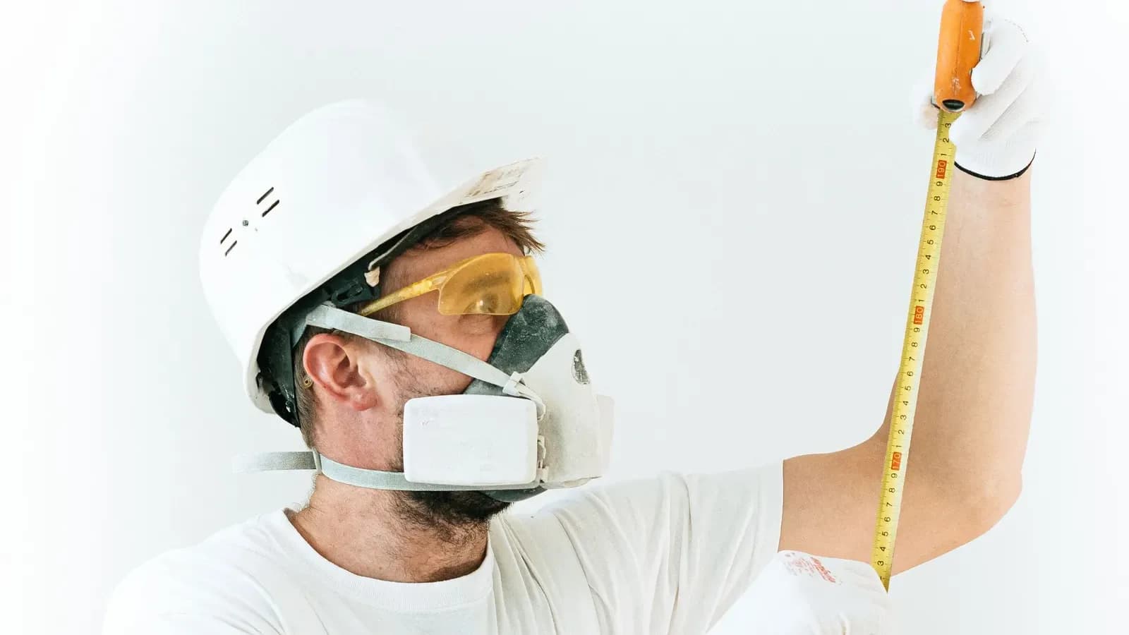 A worker in a white hard hat, dust mask and yellow safety glasses reading a yellow tape measure held up beside his face