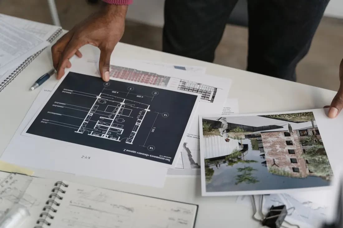 Two hands holding a printed floor plan over a desk of drawings, with a pen and notebook beside it