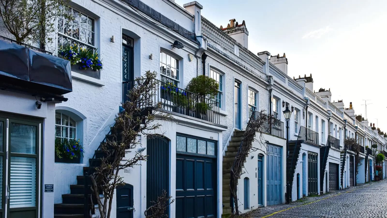 A London mews of white-painted two-storey houses with garage doors and external stairs along a cobbled street