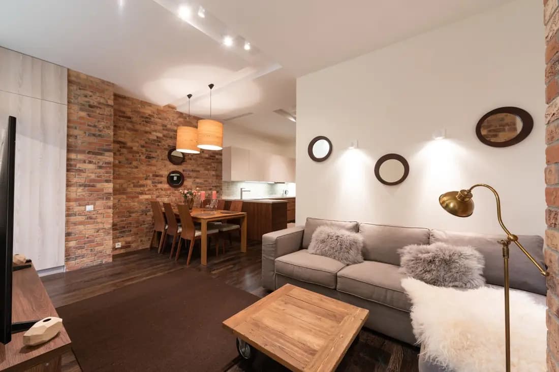 An open-plan living room with an exposed brick wall, a sofa and a dining table under pendant lights