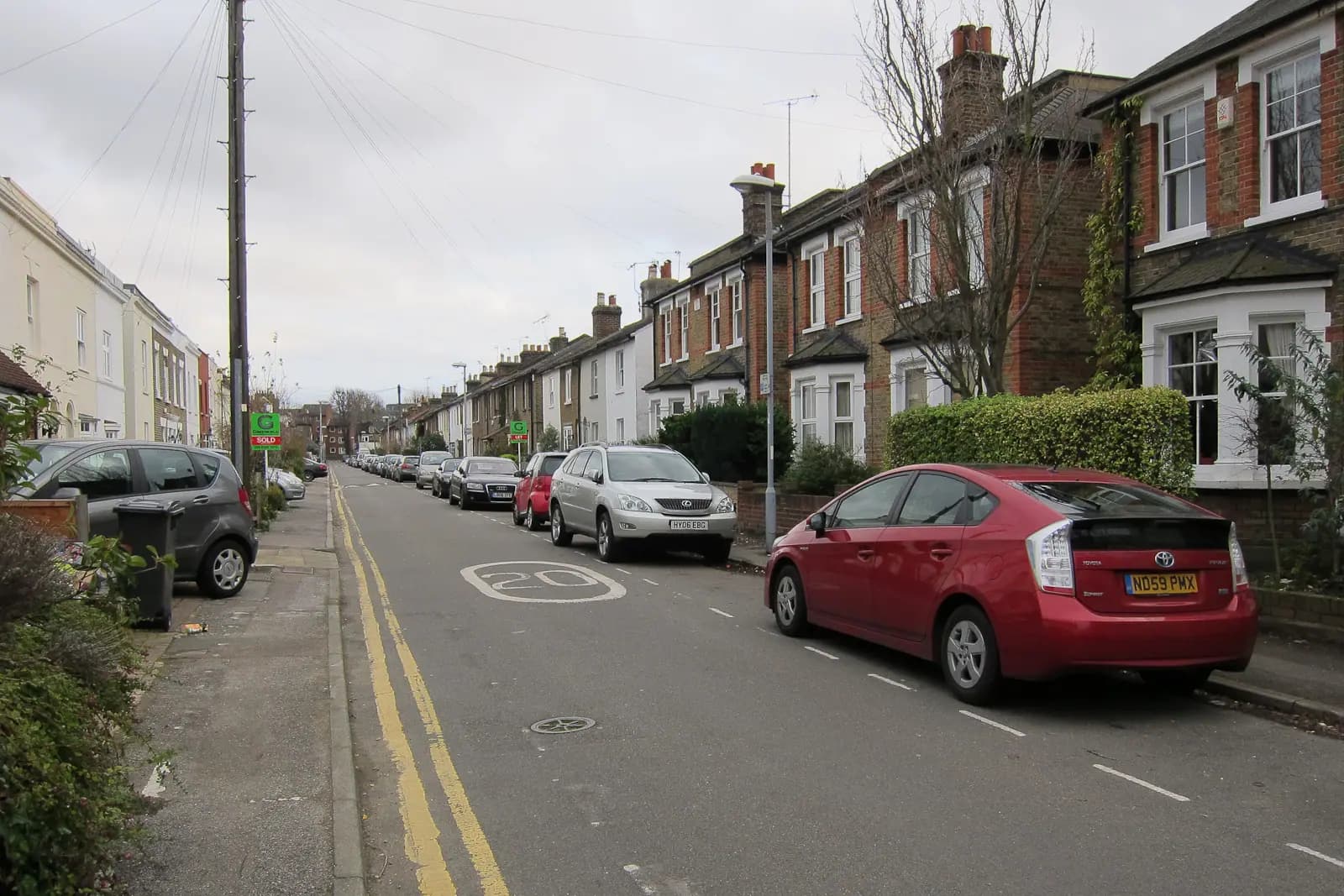 A residential street in Surbiton lined with brick Victorian terraced houses with white bay windows, a red car parked on the right and a 20 mph road marking