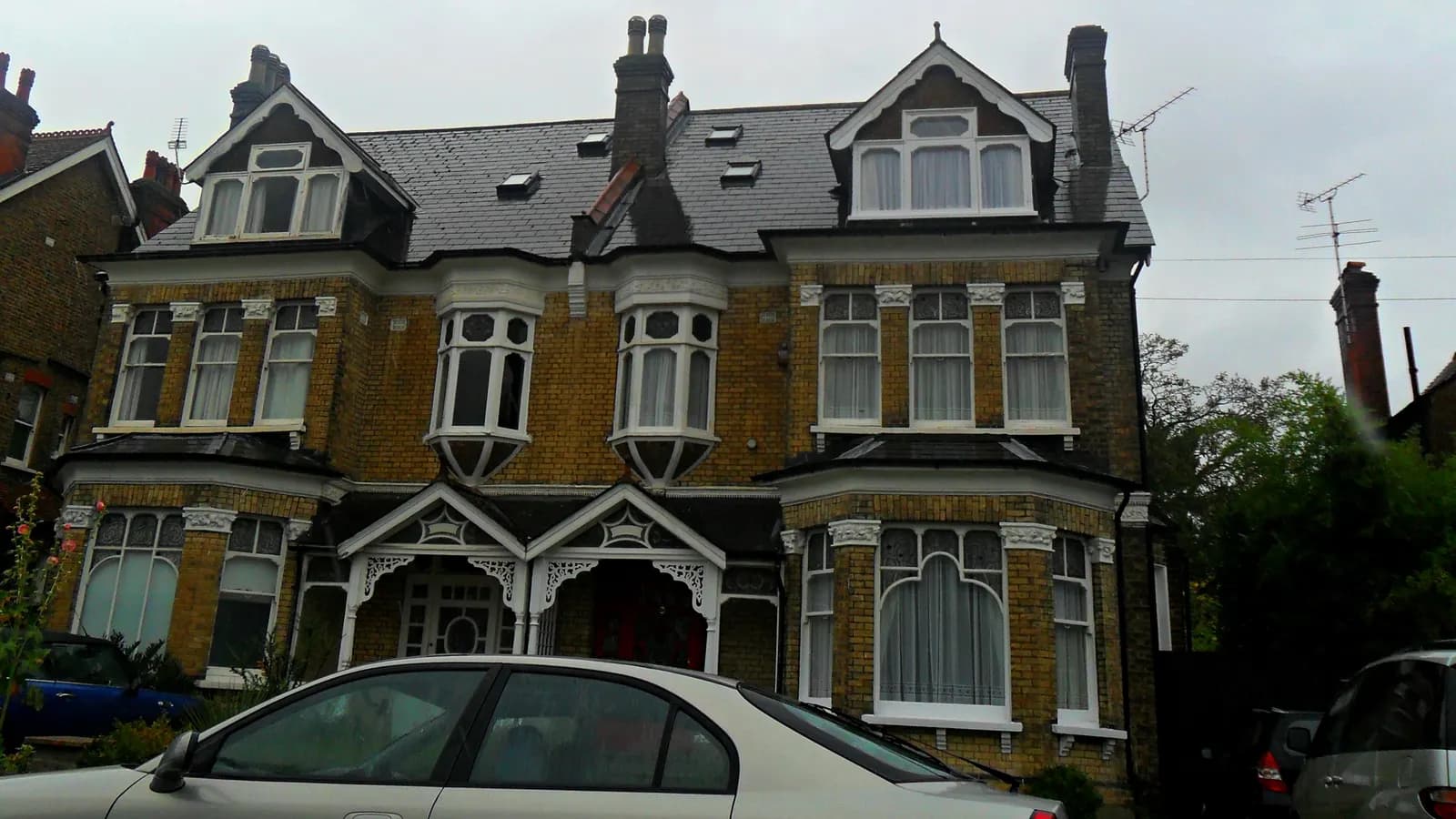 A large Victorian yellow-brick house in Sutton with carved white porches, bay windows and dormers, cars parked in front