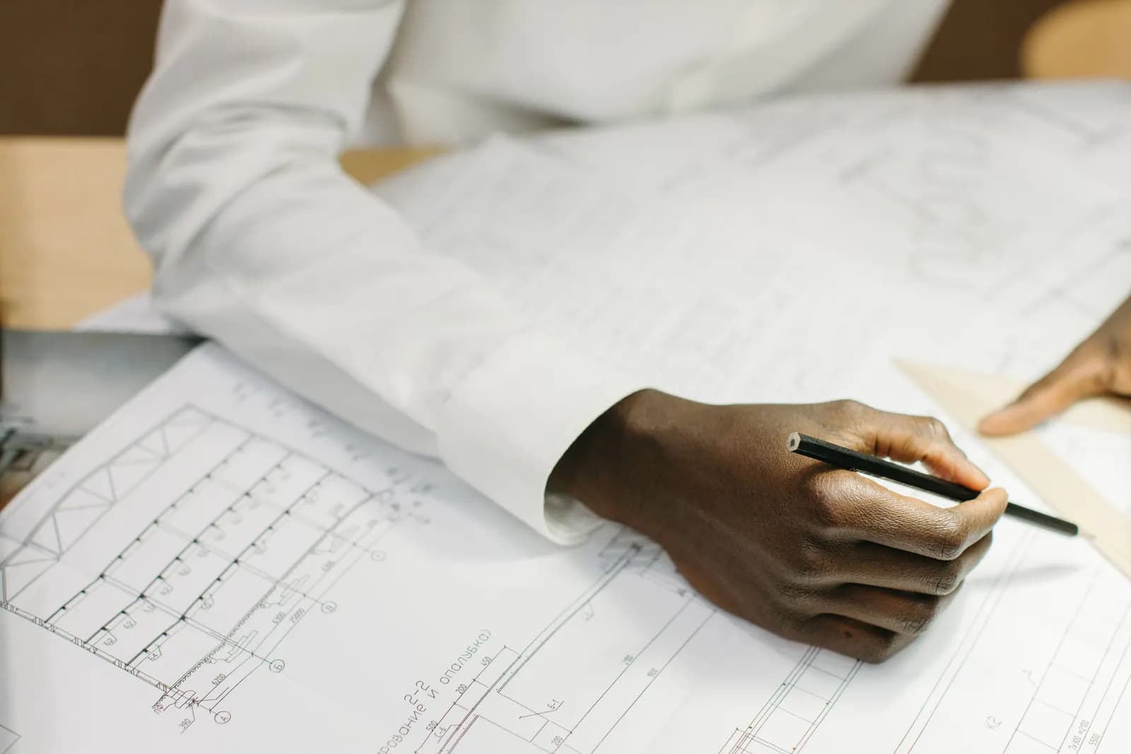 A hand holding a pencil over technical building drawings, with the edge of a wooden set square at the right of the sheet