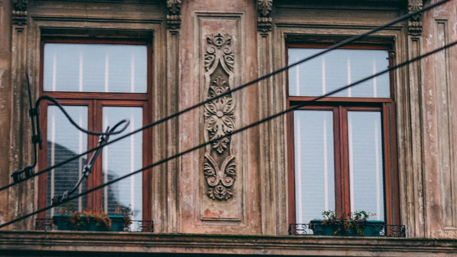 Close-up of an old stucco facade with two tall red-framed windows, carved floral relief between them and cables strung across the front