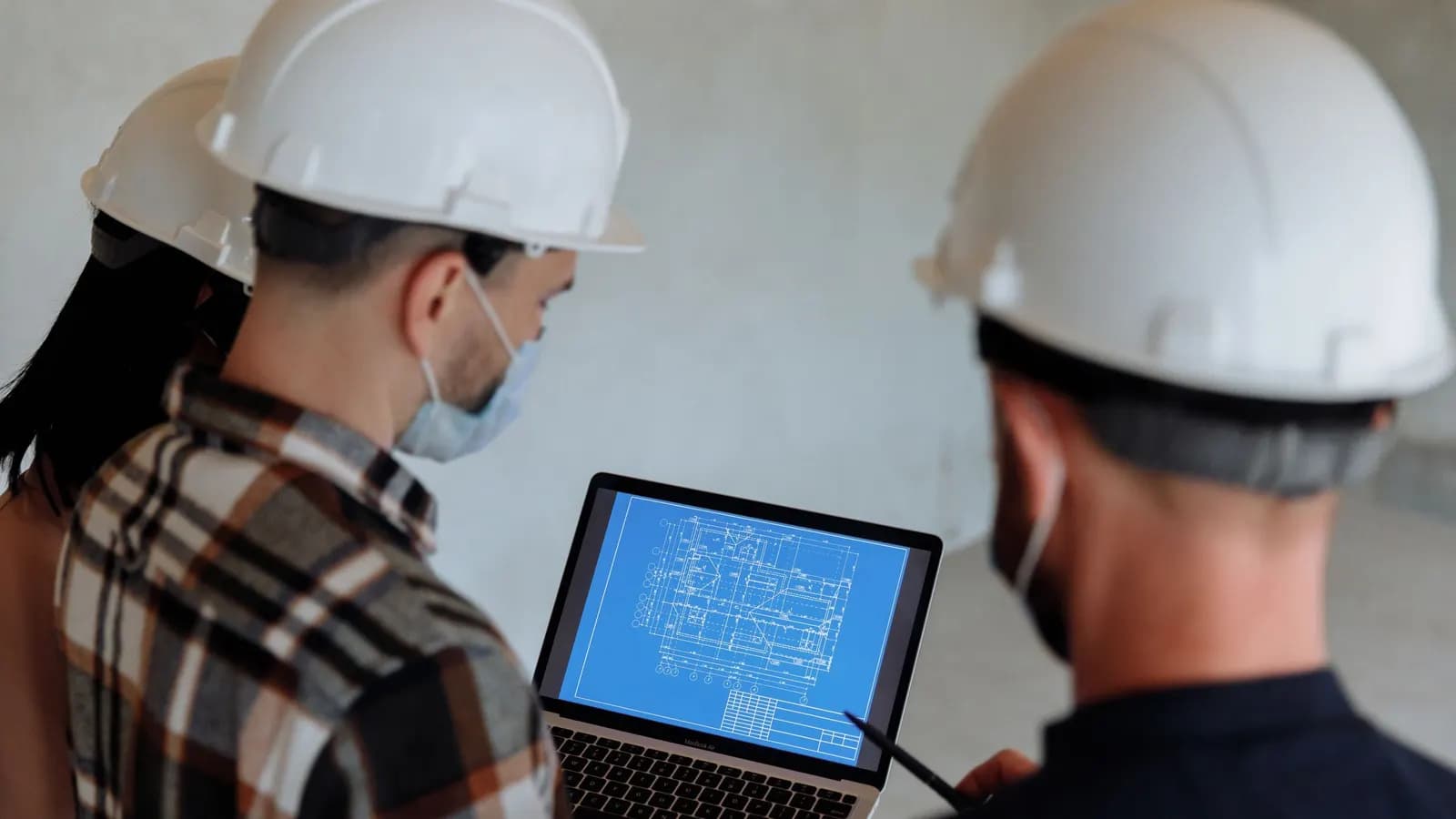 Three people in white hard hats and face masks looking at a building plan on a laptop screen