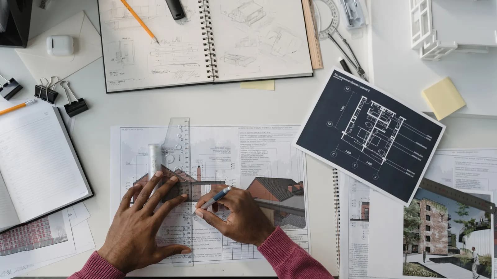 Hands drawing along a ruler on a plan, with a sketchbook, a printed floor plan and a small white model on the desk
