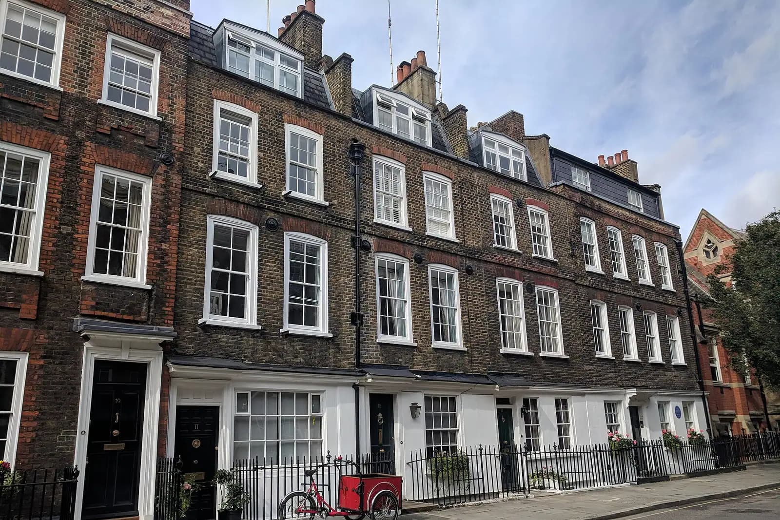 A terrace of dark Georgian brick houses with white sash windows, black railings and a red cargo bike outside on a London street