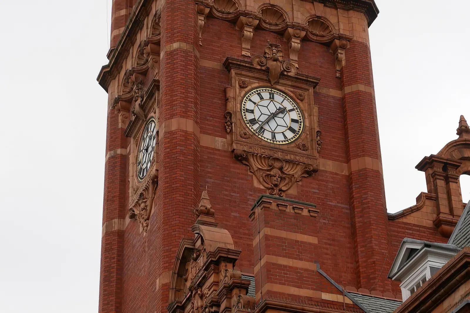 A red-brick clock tower with terracotta decoration and a white clock face with black Roman numerals, against a pale sky