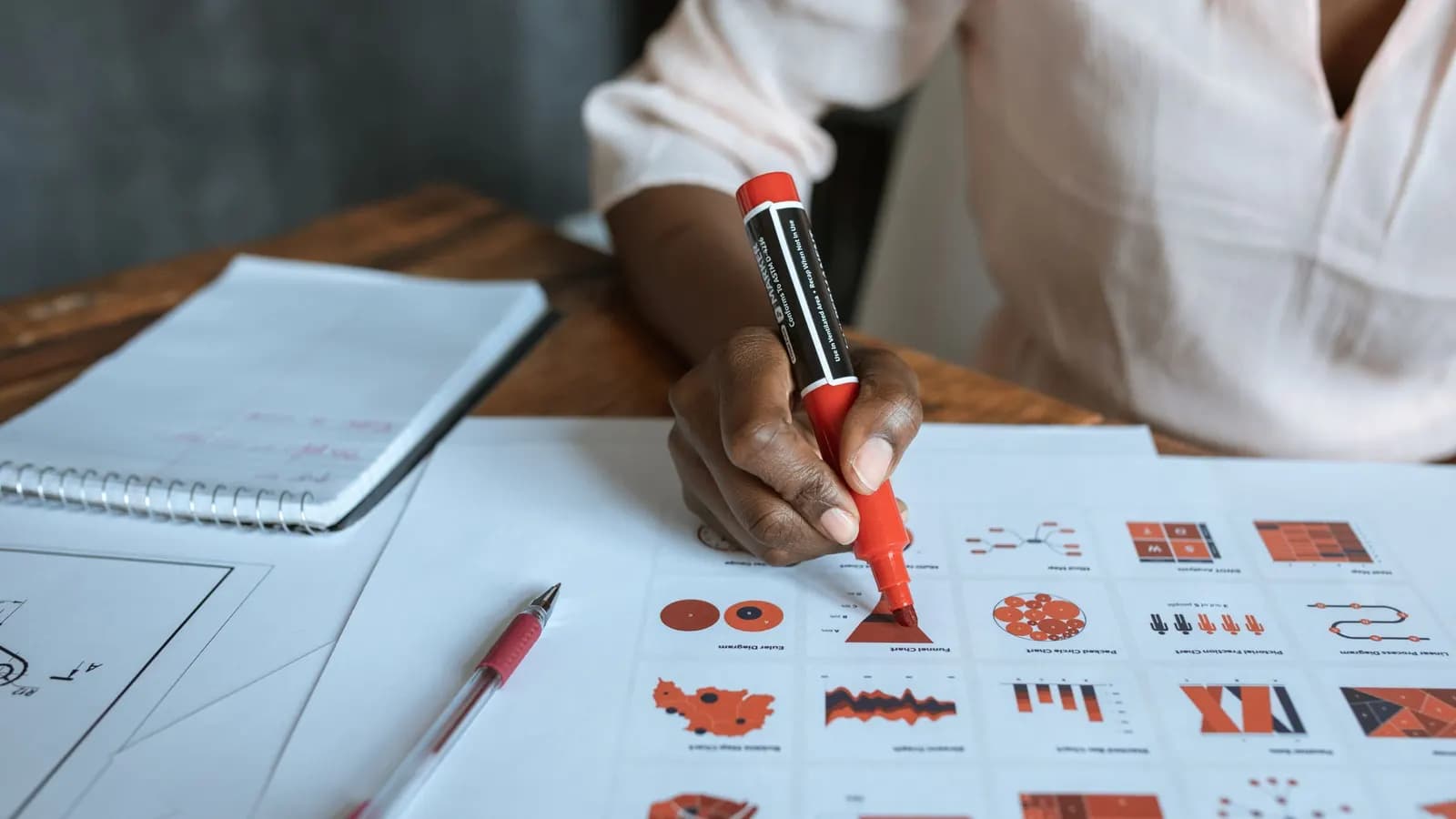 A hand marking up printed charts with a red marker, beside a notepad, a pen and part of a technical drawing