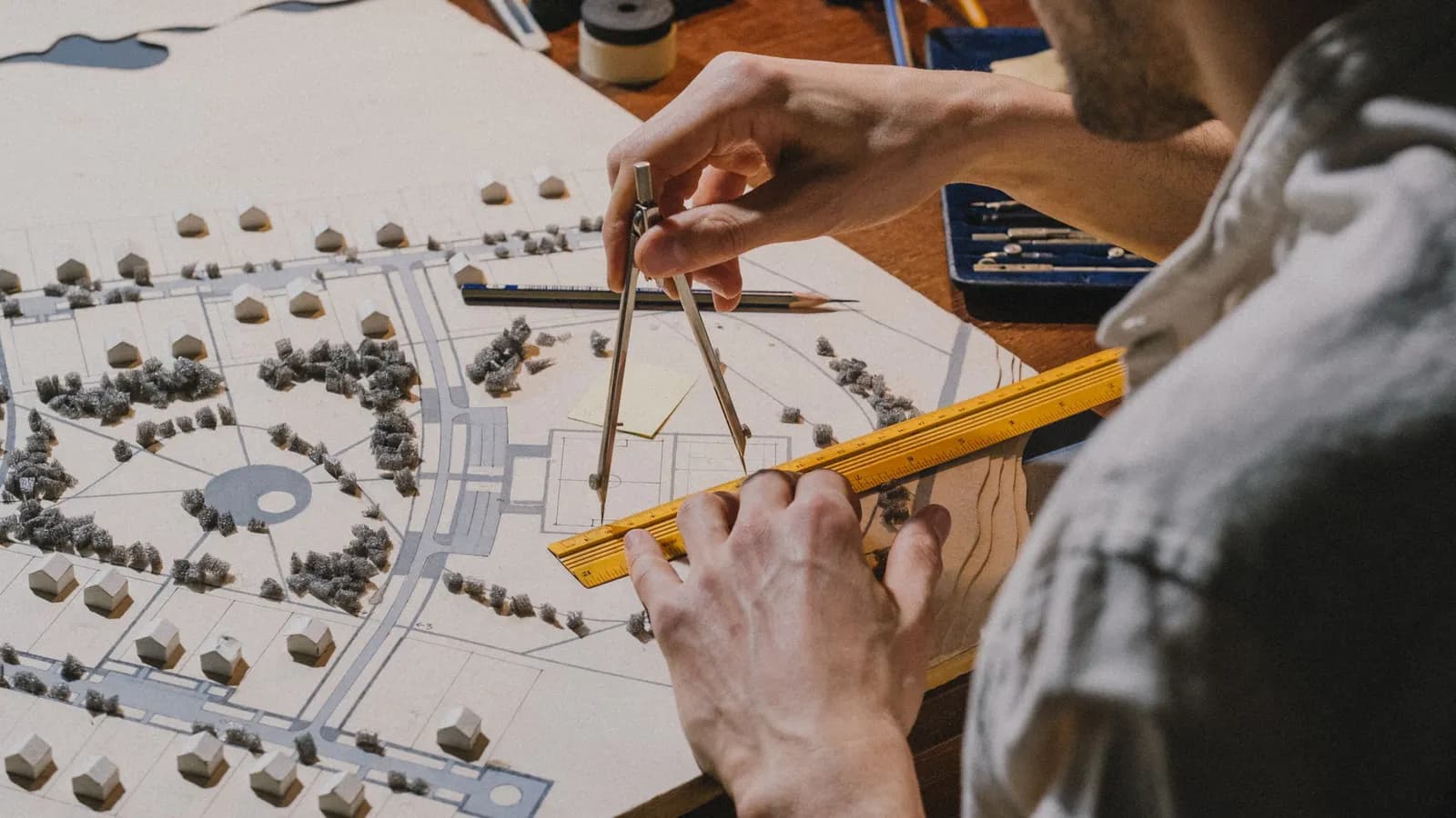 A person using dividers and a yellow ruler on a card site model with small white houses and foam trees