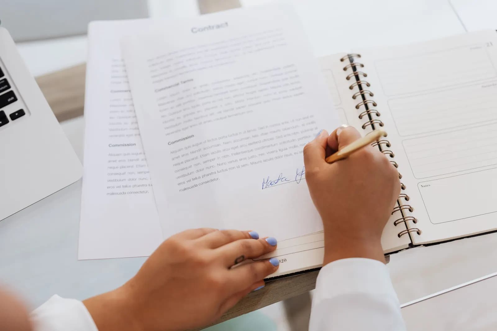 Hands signing a contract on a desk beside a spiral notebook and the edge of a laptop