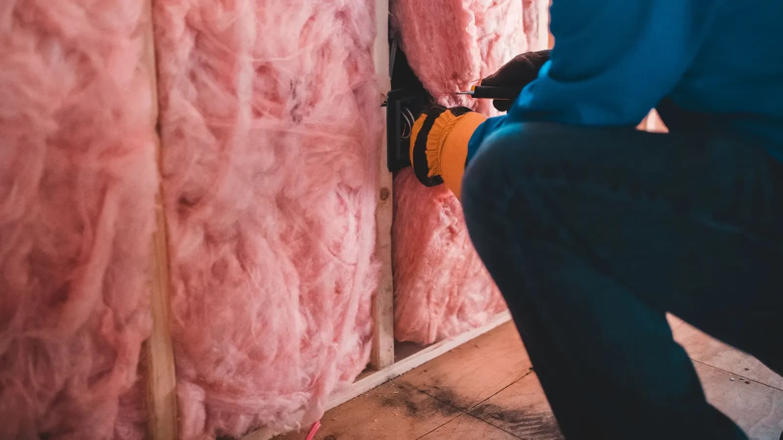 A worker in a blue top and gloves crouching beside pink insulation fitted between timber studs