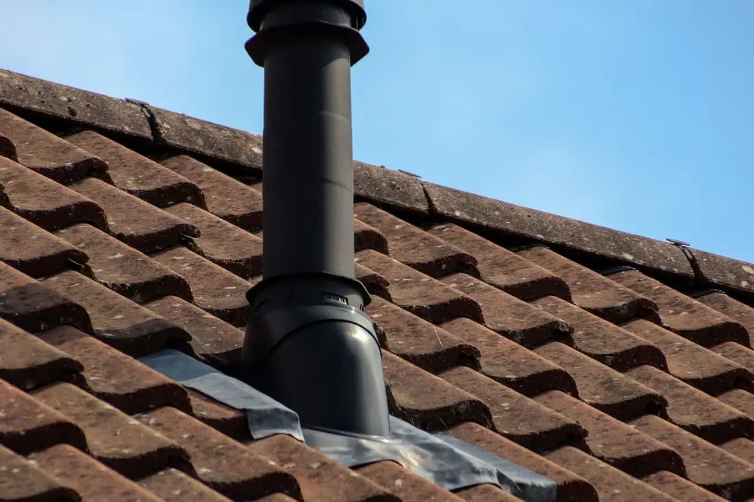 Clay roof tiles on a pitched house roof with a black flue pipe passing through, against a blue sky