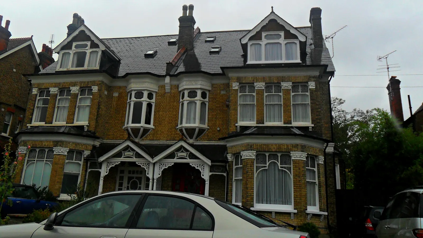 A large Victorian yellow-brick house in Sutton with carved white porches, bay windows and dormers, cars parked in front