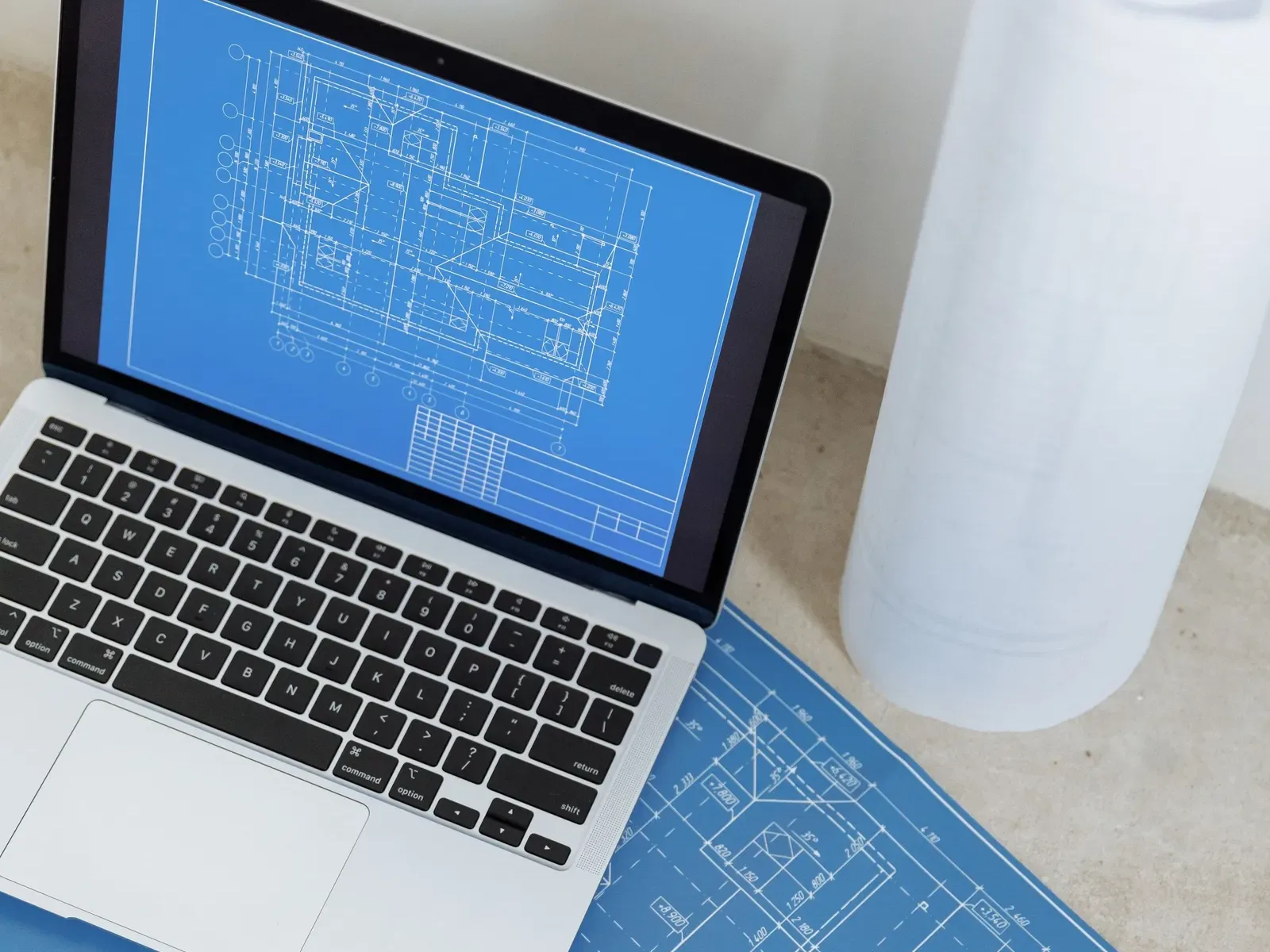 A laptop showing a building plan beside a printed blue construction drawing and a roll of paper