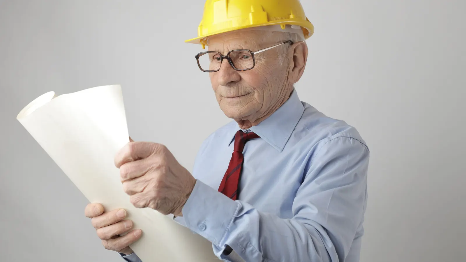An older man in a yellow hard hat and blue shirt reading a rolled-up plan
