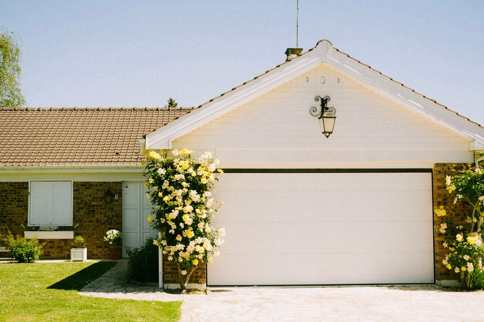 A single-storey brick house with a large white garage door, a paved drive and climbing yellow roses