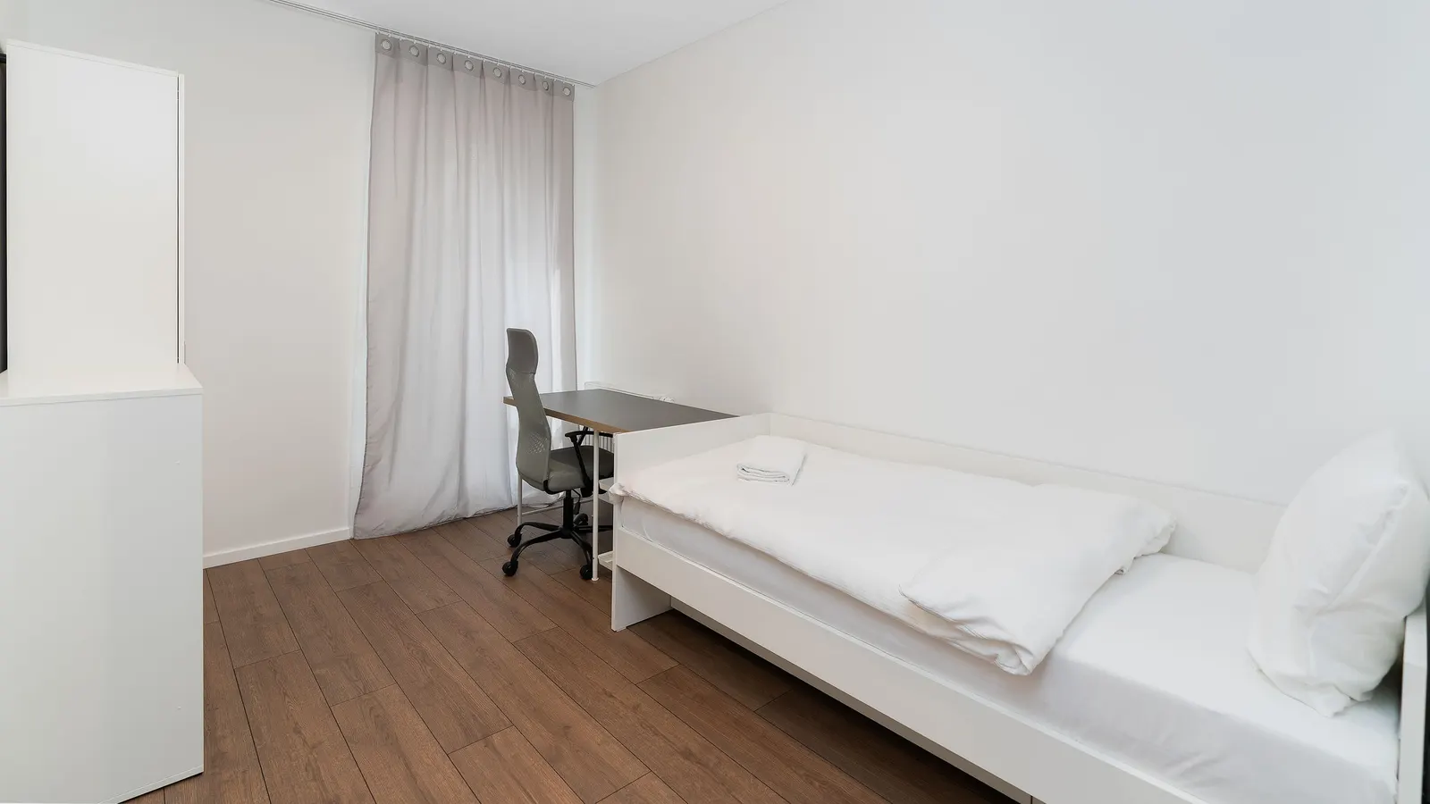 A plain single bedroom with a white-covered single bed, a desk and grey office chair beside a pale curtain, on a wood-effect floor