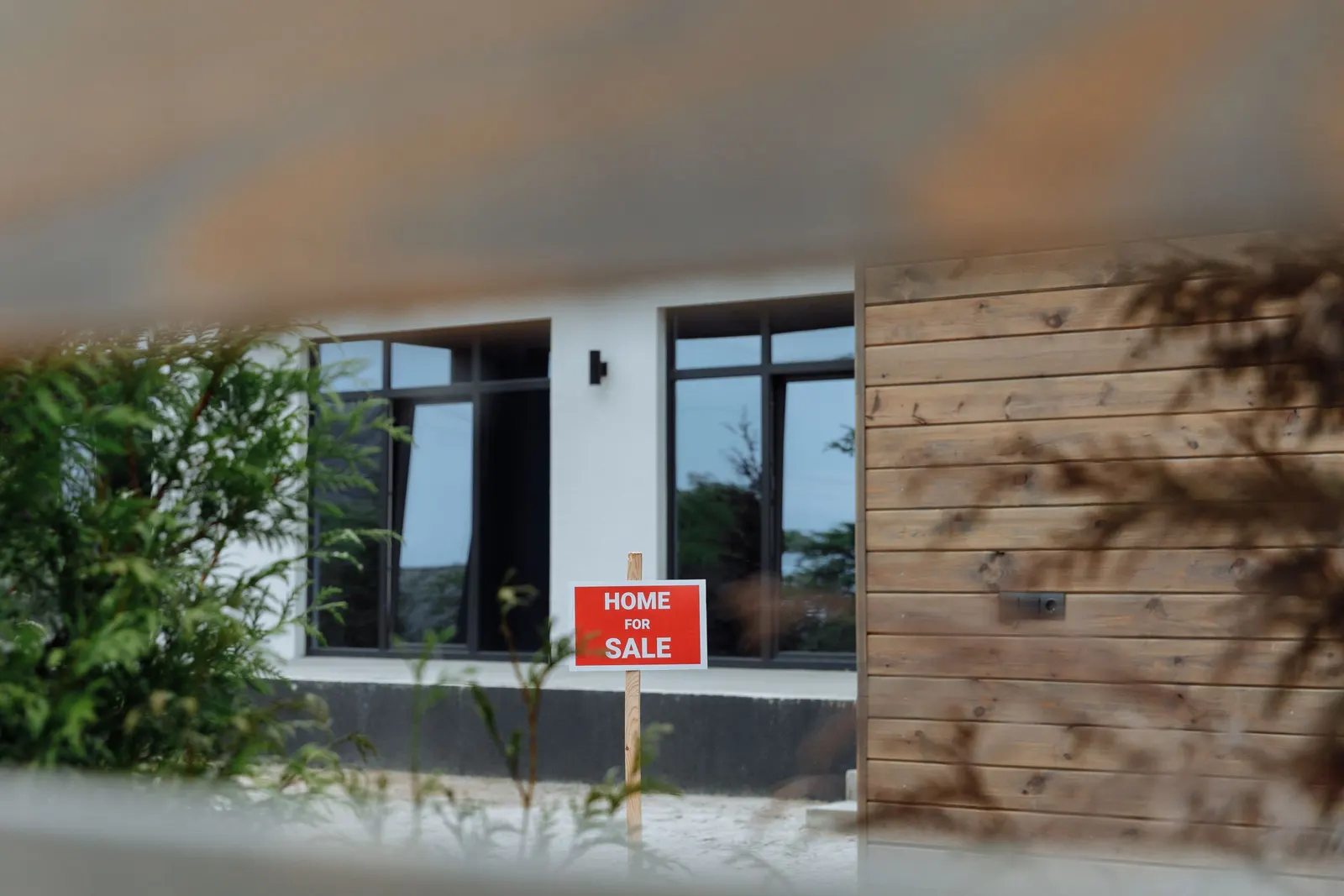 A house for sale sign in the front garden of a modern house with large dark-framed windows, seen through leaves