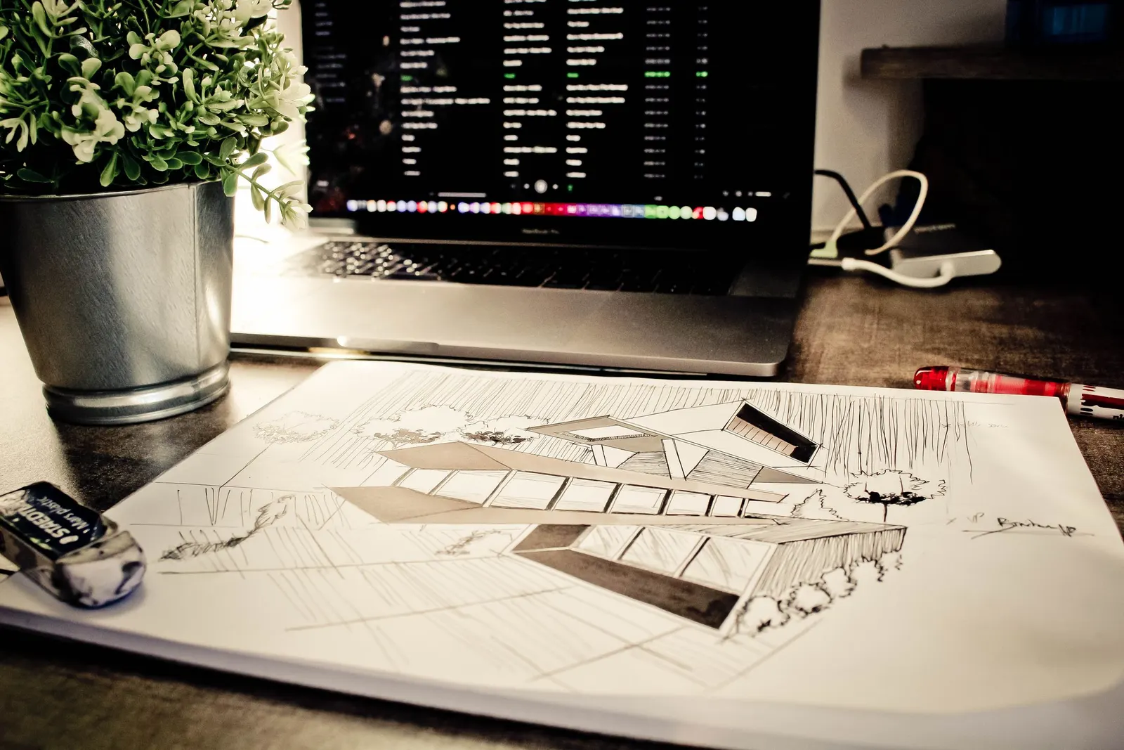 A pen-and-ink sketch of a modern building on a sheet of paper, with a laptop, a potted plant and an eraser on the desk