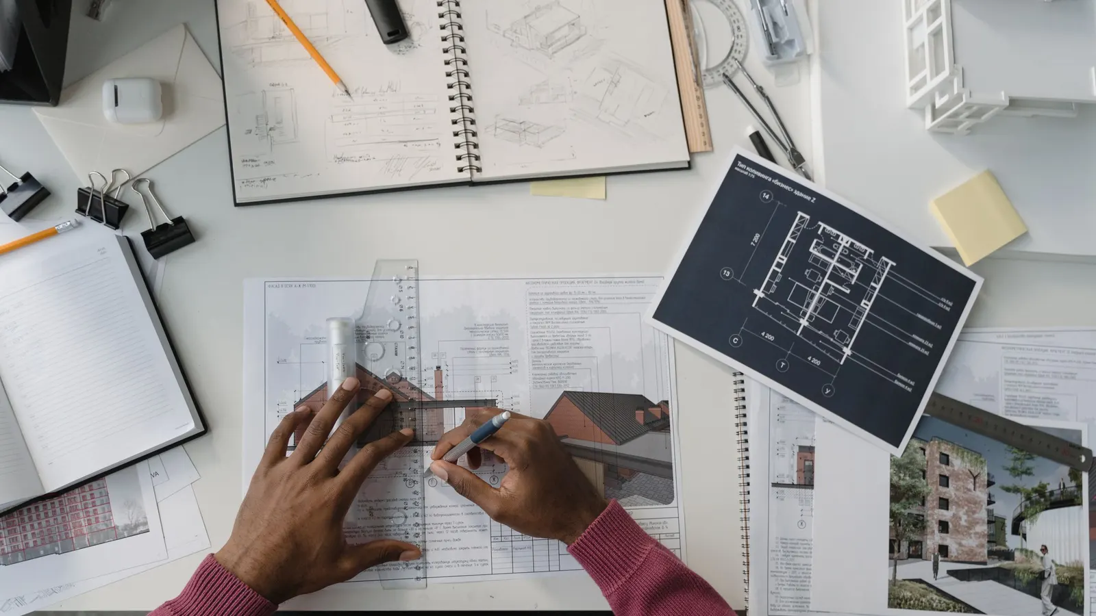 Hands drawing along a ruler on a plan, with a sketchbook, a printed floor plan and a small white model on the desk