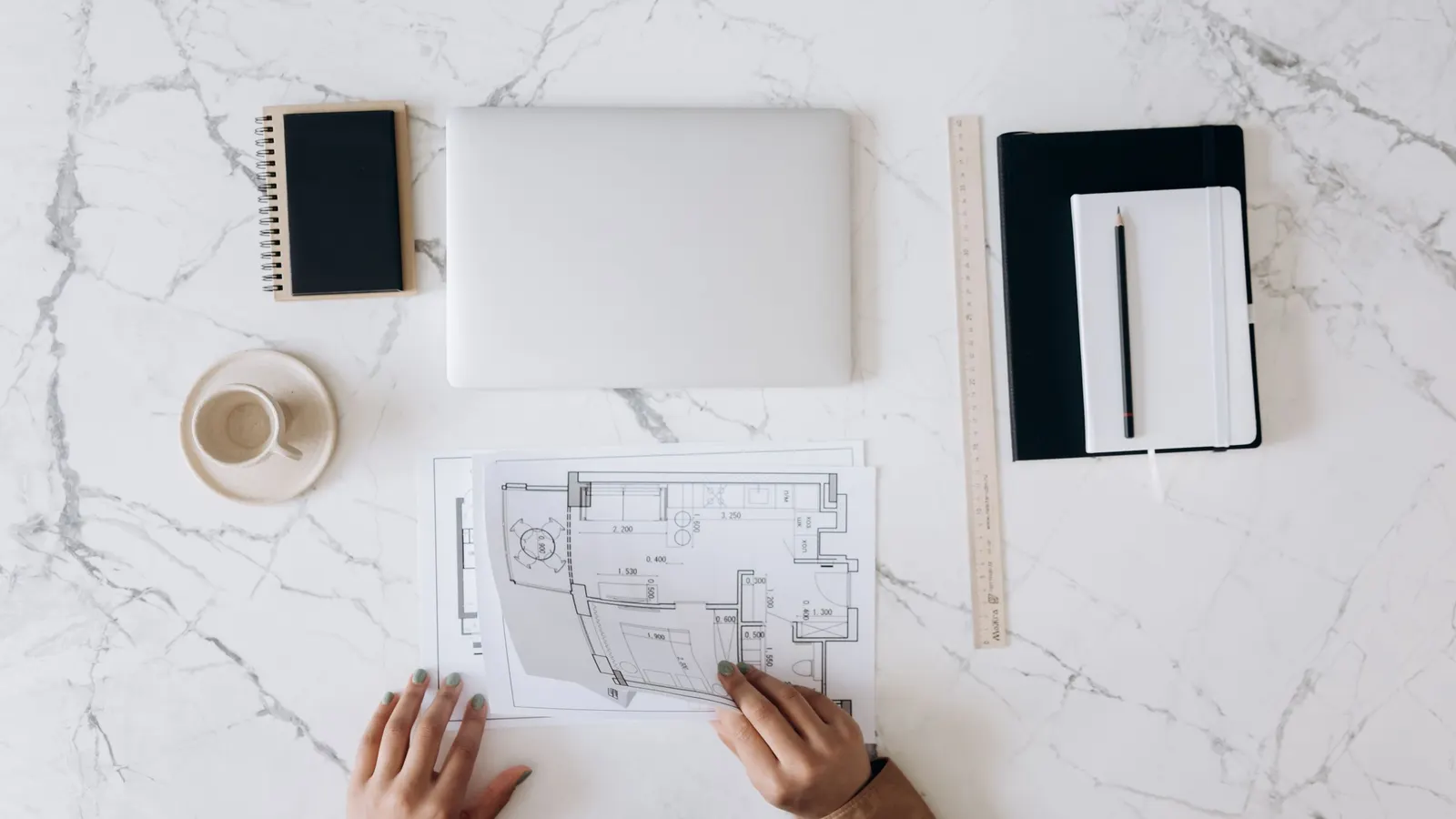 A top view of a white marble desk with a closed laptop, notebooks, a ruler, a coffee cup and two hands turning a floor plan