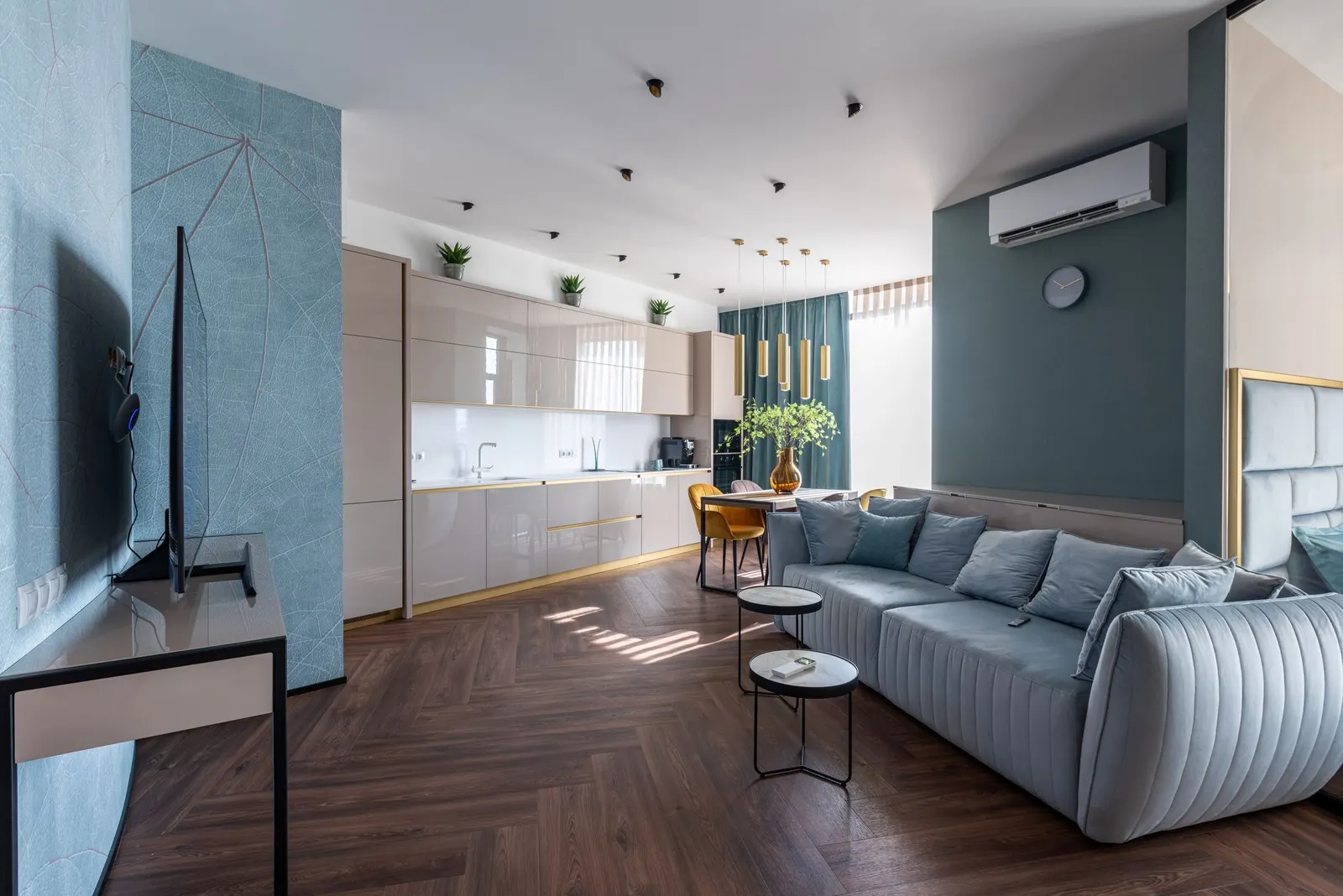 An open-plan living space with a pale blue sofa, a kitchen run along one wall and a dining table by the window