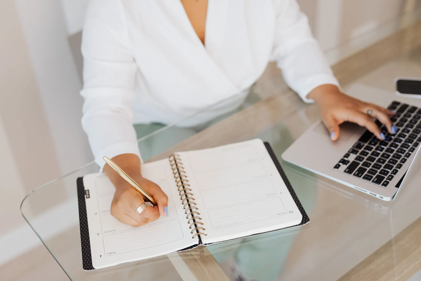 A person writing in a diary with a gold pen while typing on a laptop at a glass desk