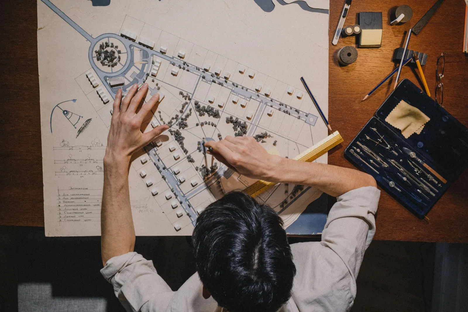 A person seen from above placing small white model buildings on a hand-drawn site plan, beside a case of compasses and pencils