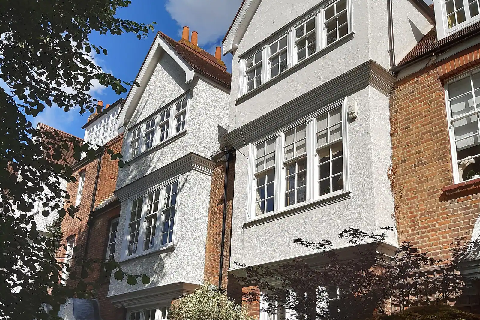Two tall white roughcast houses with gabled bays and white sash windows between red brick walls, with a Japanese maple in front