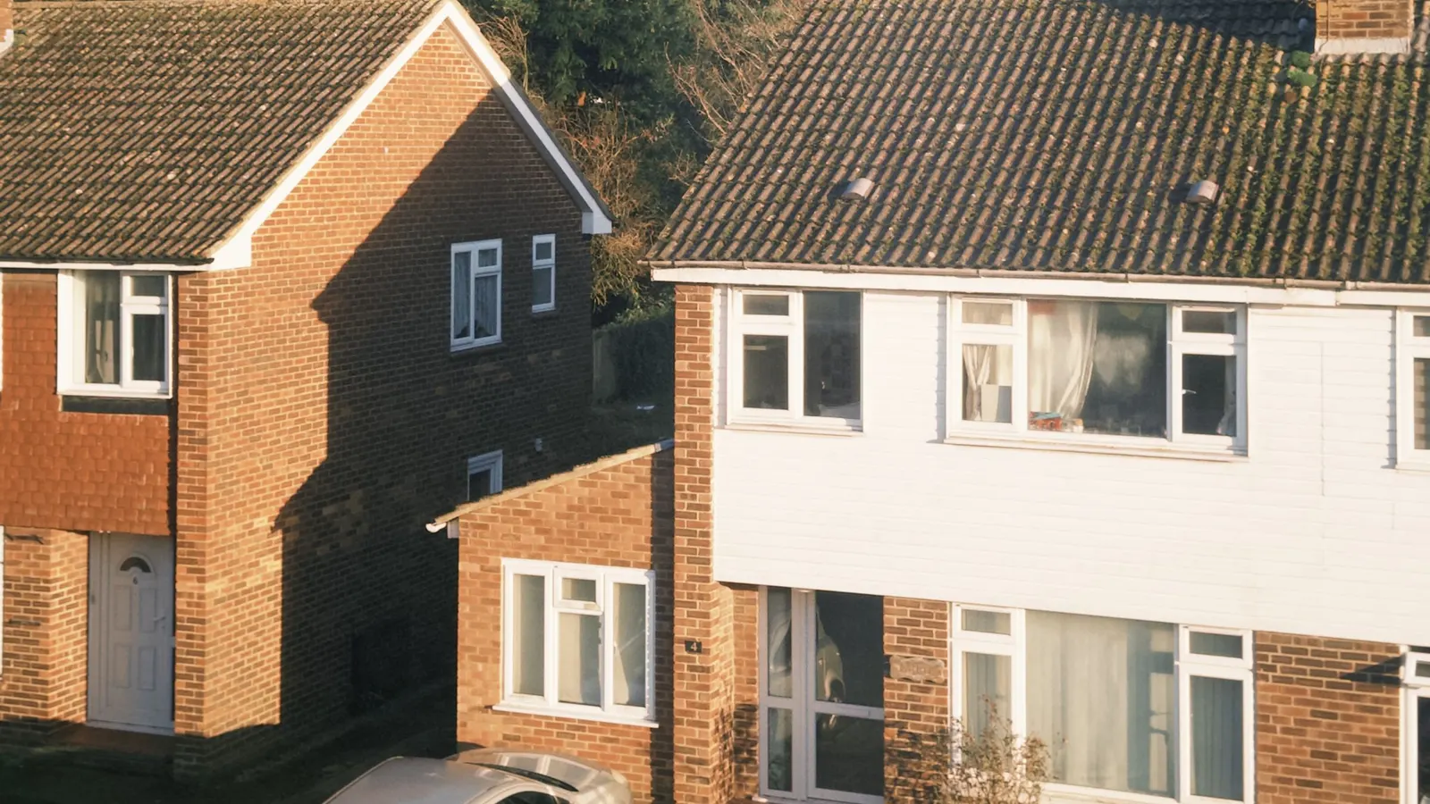 A brick detached house beside a semi-detached pair with white weatherboarding and a single-storey side wing, in low evening sun