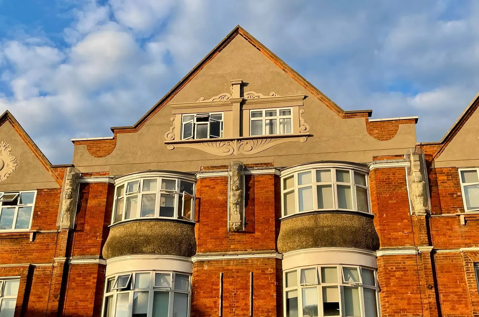 The upper floors of a red brick building with white-framed curved bay windows, carved stone figures between them and a plastered gable under a blue sky