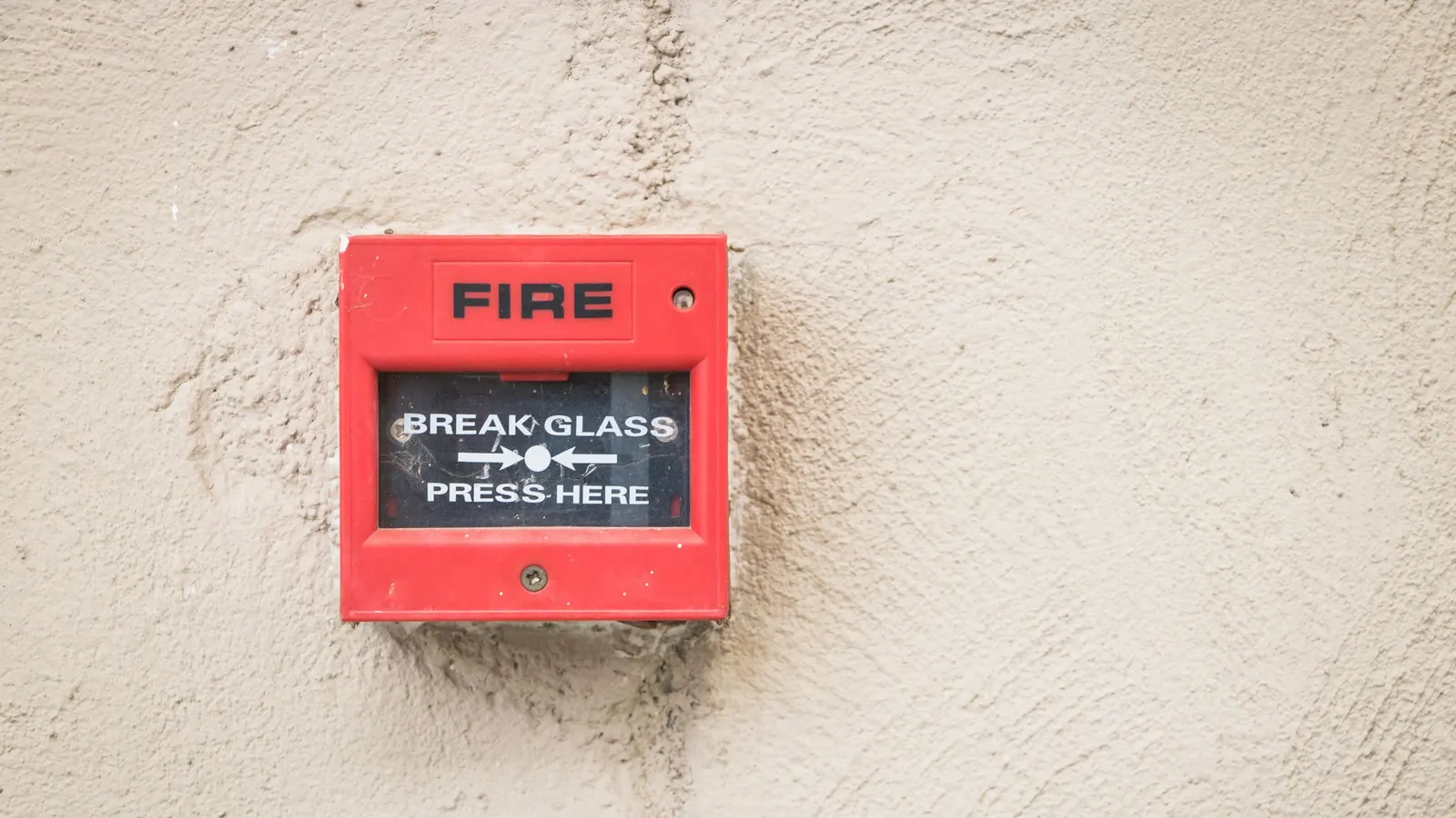 A red break-glass fire alarm call point fixed to a rough cream-coloured wall