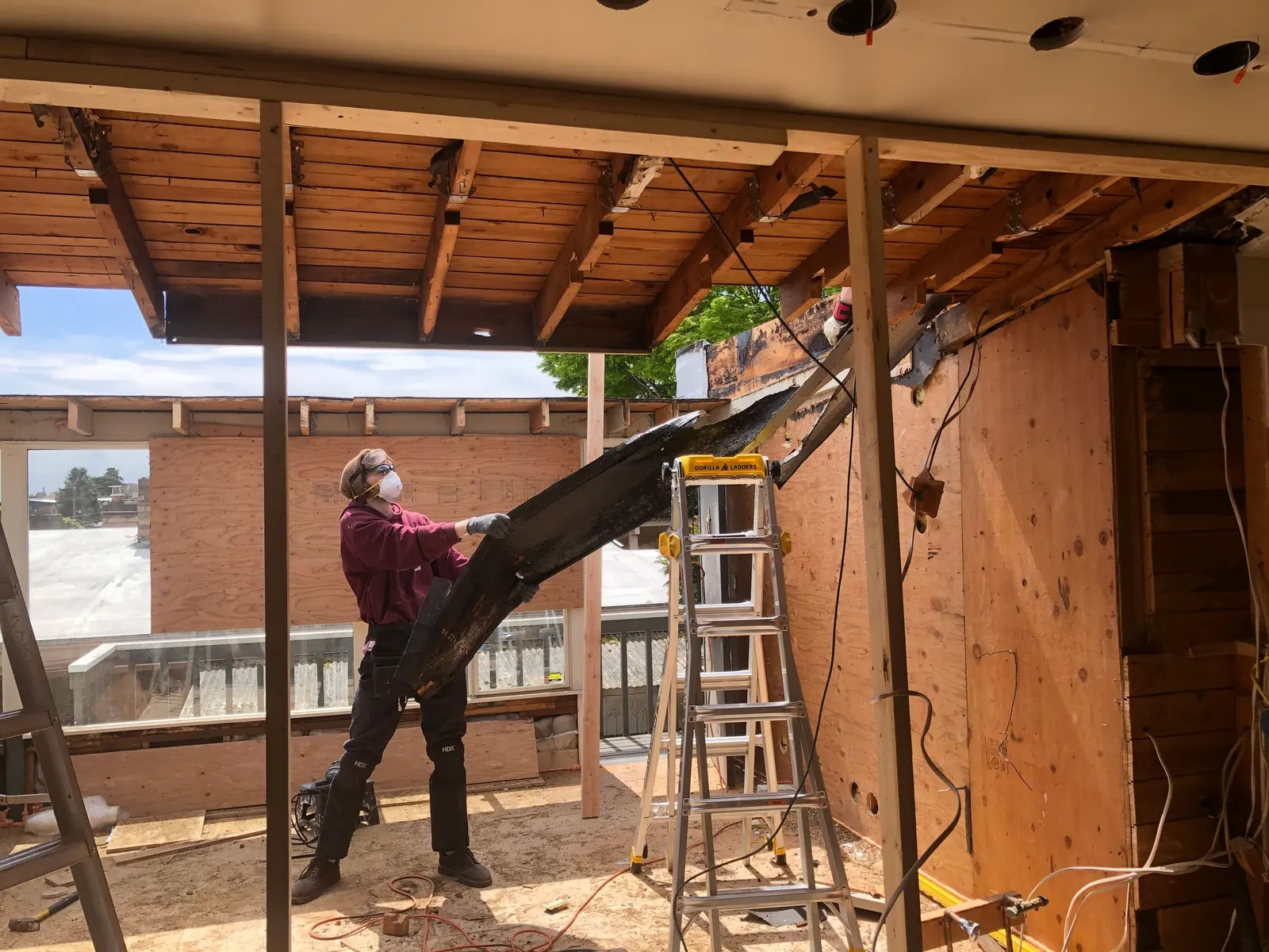 A worker in a dust mask and safety glasses lifting a charred timber out of an opened-up roof and ceiling beside a ladder