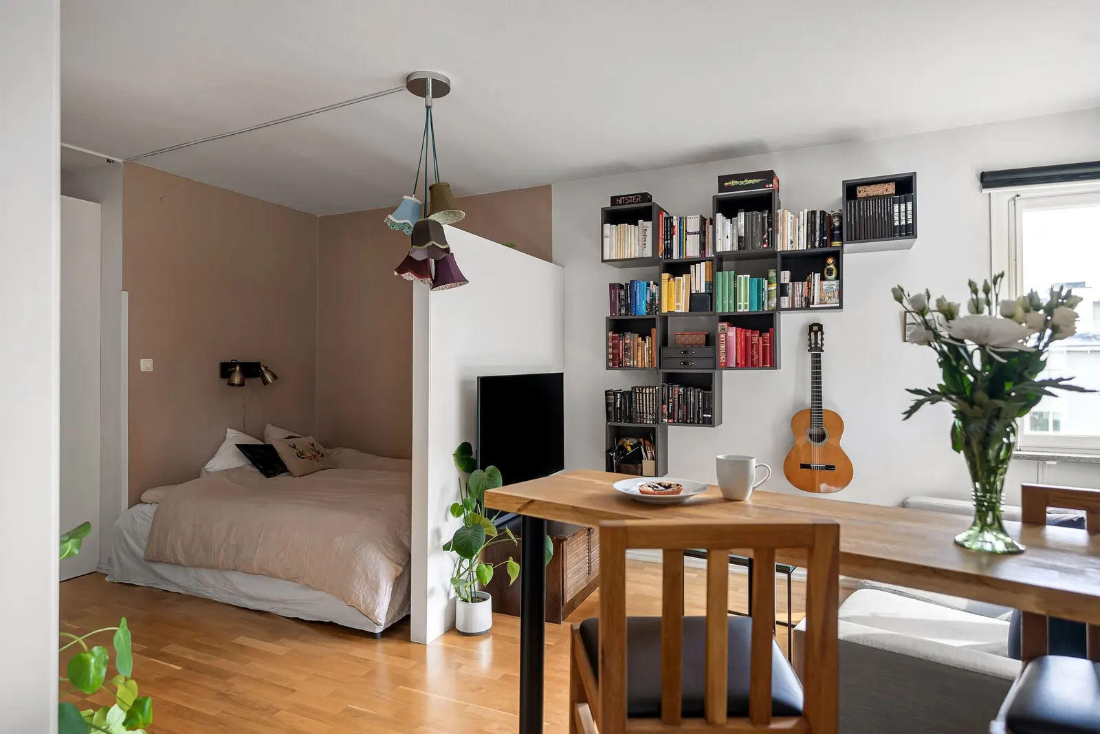 An open-plan studio with a bed in an alcove behind a low partition, a wall of shelves with books, a guitar and a wooden dining table