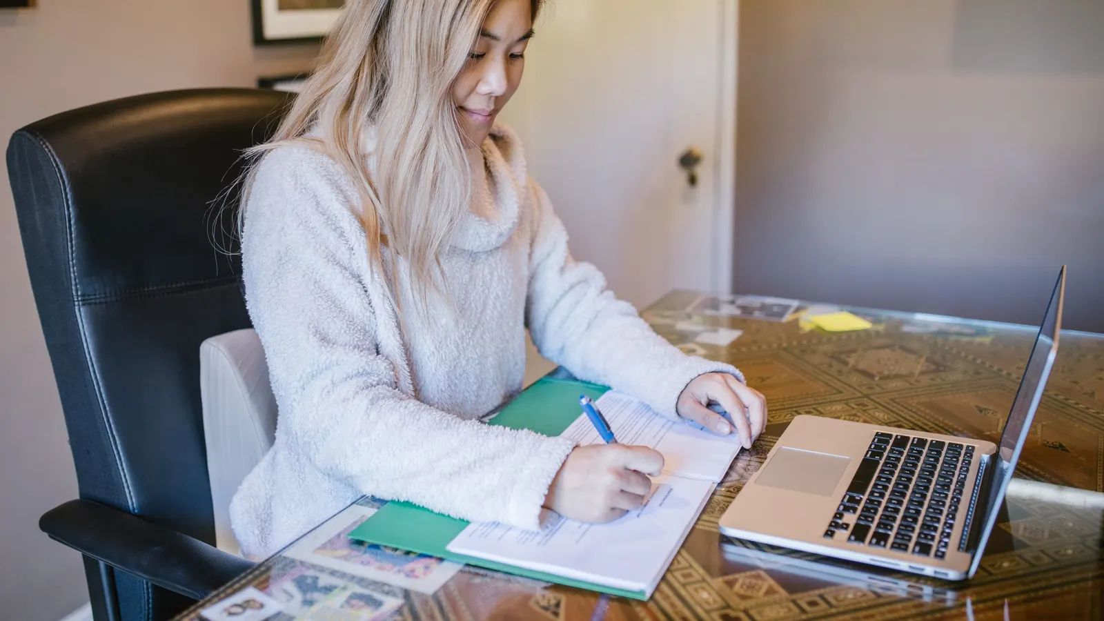A woman at a desk filling in a form with a pen, beside a laptop and a green folder