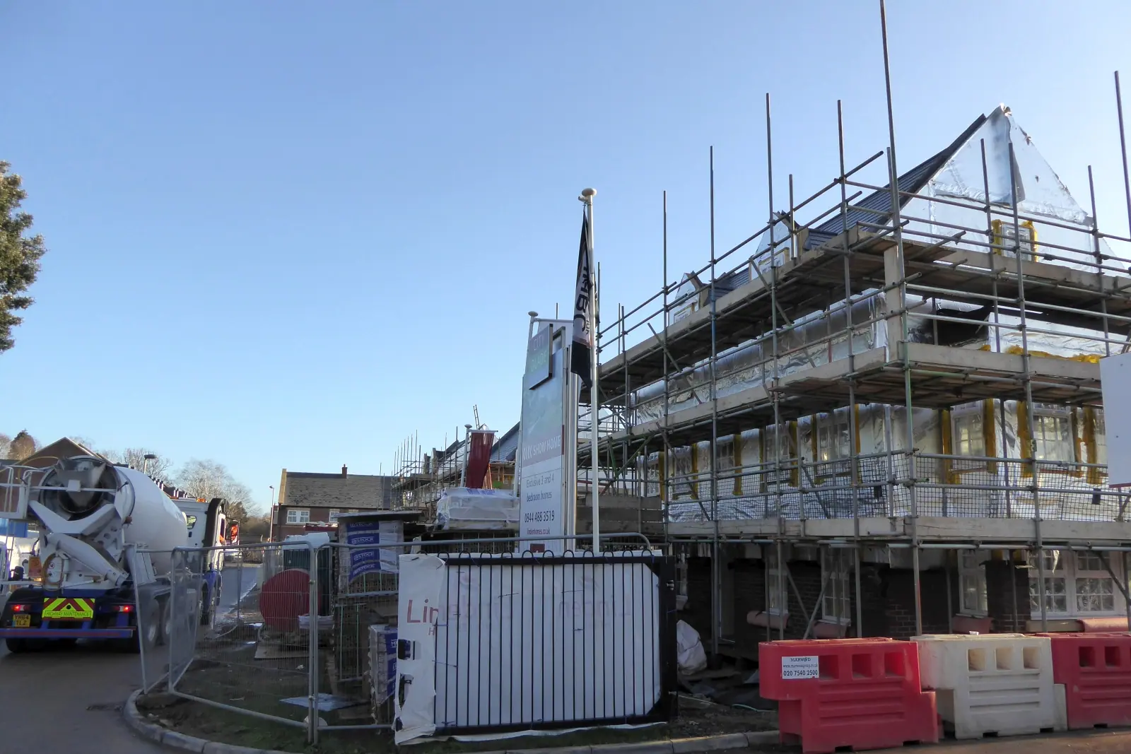 A new-build house under construction wrapped in scaffolding with a concrete mixer lorry, site fencing and barriers beside it
