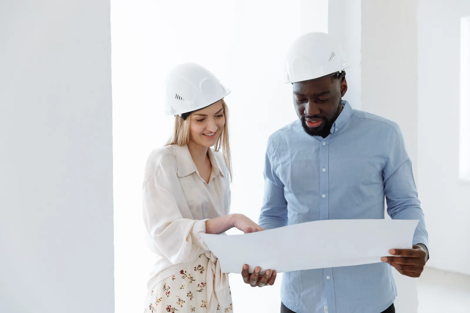 A woman and a man in white hard hats looking at a large printed plan in a bright empty room