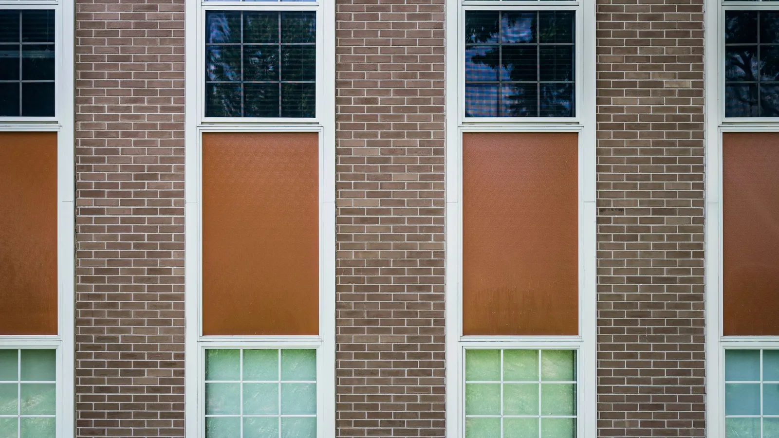 A brown brick wall with tall white-framed windows, some filled with orange and green panels